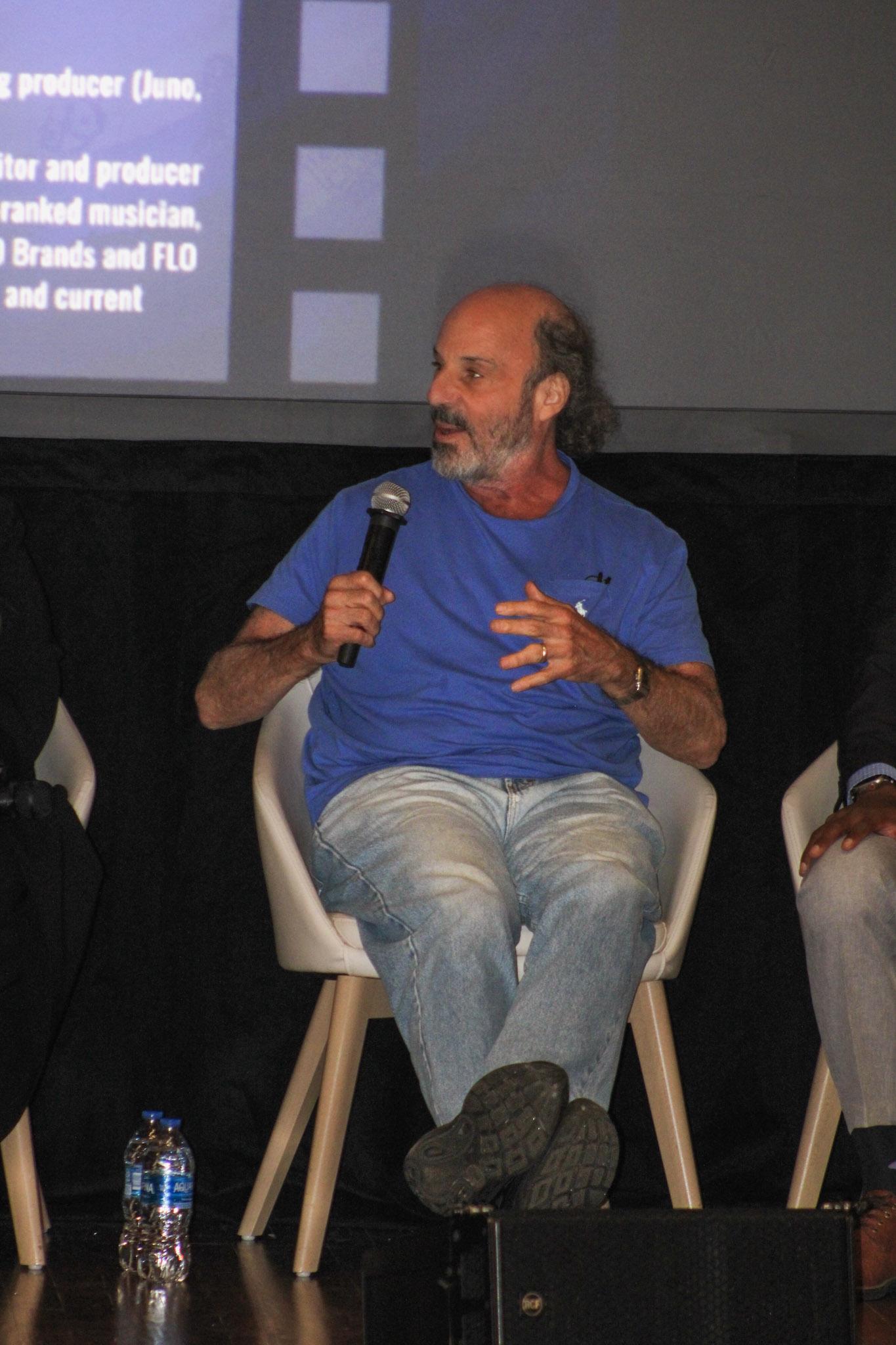 A man sitting on a chair on a stage. He is holding a microphone and is in mid sentence