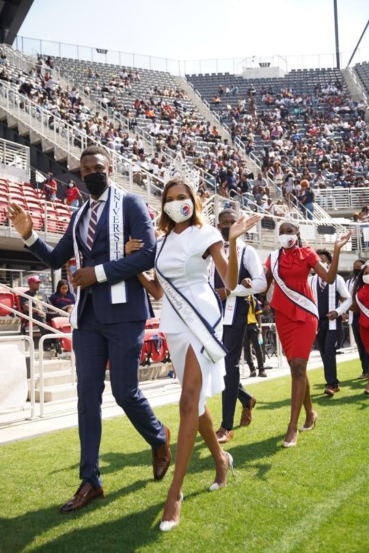 Howard's Royal Court members walking across the Howard University's football field. There is a young man and woman arm in arm, who are waving. There are wearing face masks