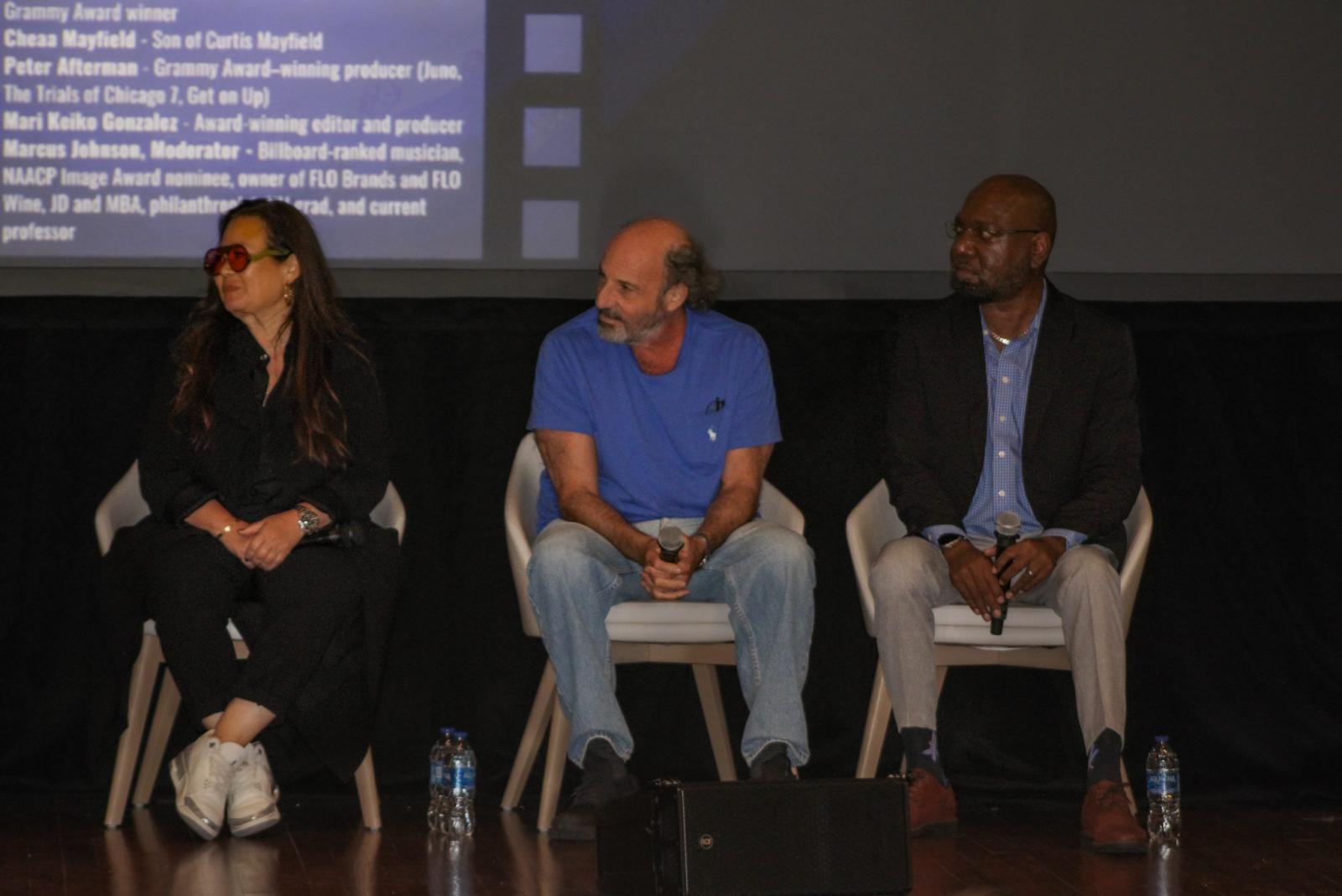 Three people sitting on a stage. The person in the middle and the person on the right are holding microphones.