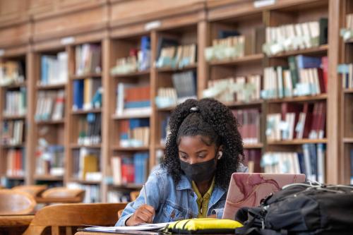 Student wearing a face mask studies with laptop and notebooks in library