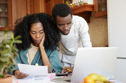 Young african family paying utility bills online using laptop computer unhappy female sitting table analyzing sheet paper her hand calculating domestic expenses together with her husband
