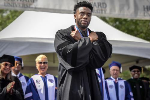 Chadwick Boseman doing the Wakanda Salute pose on stage. He has his arms crossed on his chest. There are people standing behind him