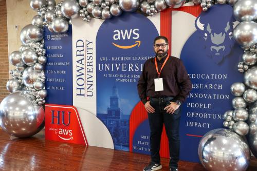 Man in black shirt and slacks stands in front of Amazon Web Services and Howard University step and repeat