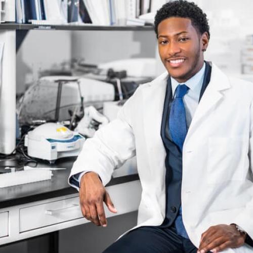 A smiling young man in a lab coat surrounded by lab equipment.