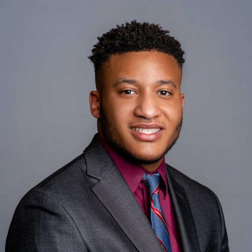 Kaie Hall wearing a dark grey suit jacket with a dark red button down sir shirt and a red and blue patterned tie