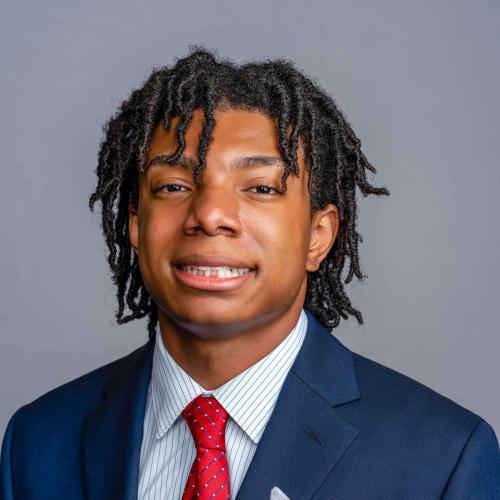 Esau Hutcherson wearing a navy blue suit jacket with a white pinstripe button down shirt and a red and blue striped tie 