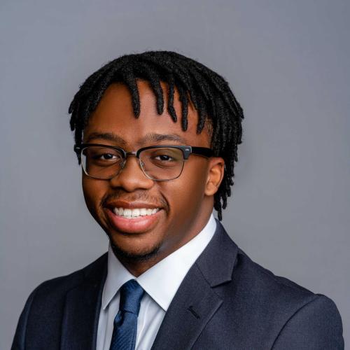 Myles Mackie wearing a dark navy blue suit jacket with a white button down shirt and a blue tie