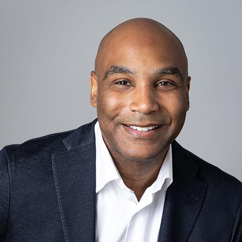 Andrew Lindsay smiles in a professional headshot wearing a navy blazer and white dress shirt against a gray background.