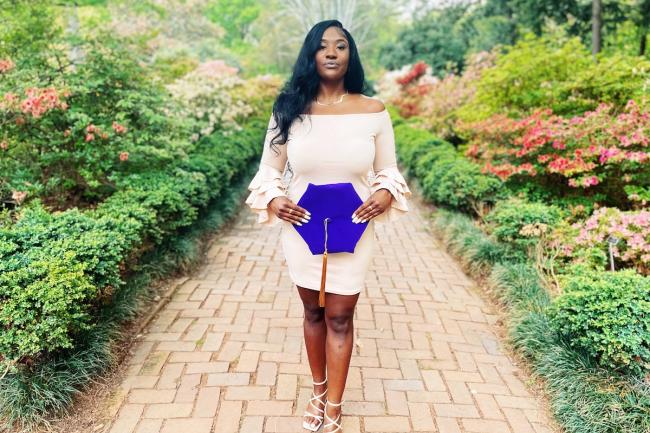 Young Black women stands on a brick path lined by flower bushes, holding a graduation cap