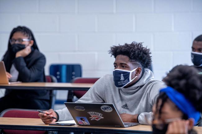 Black college student with a laptop in front of him watches his teacher during class