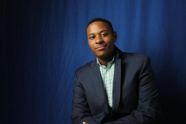 Black male student poses with arms crossed against a dark blue background