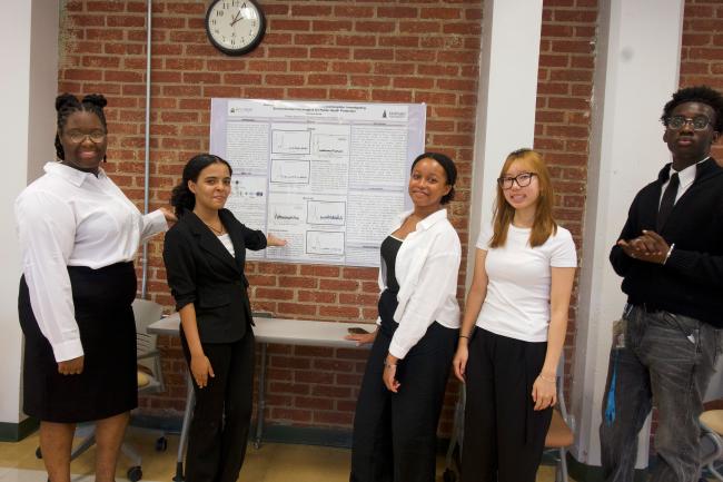 Five students smiling at the camera while standing in a classroom. Two students on the left are pointing to a poster board that displays research findings