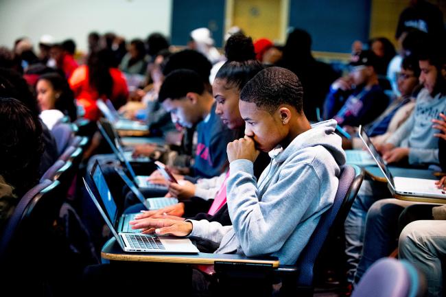 Students in a full university classroom with their laptops on the desks. The students are looking at their laptop screens intently. 