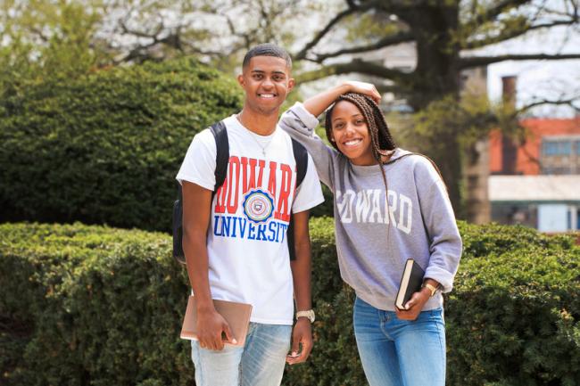 Two Howard University students standings side by side. They are wearing Howard university shirts, standing outside. In the background there are trees and grass
