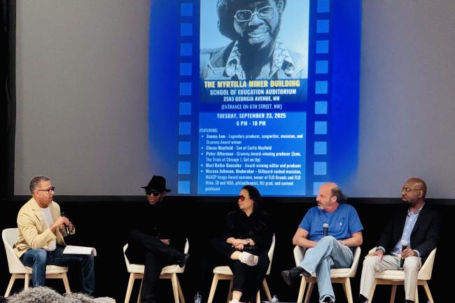 Five people sitting on a stage holding mics in their hands. There is a large projector screen behind them with an image of singer Curtis Mayfield and text