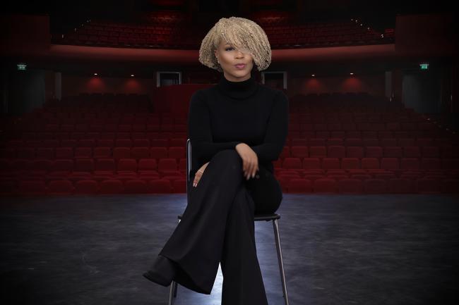 woman sitting in a chair while on a theatre stage. There are red theatre chairs behind her