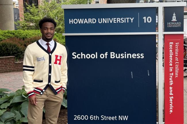 Graduating senior Joshua Taylor standS beside the Howard University School of Business sign wearing a cream colored Howard University cardigan
