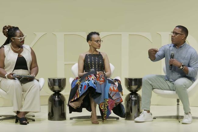 Three people sitting down on stage. Marcus johnson is on the far right, holding a mic and gesturing while talking