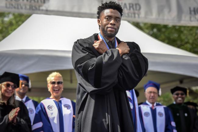 Chadwick Boseman doing the Wakanda Salute pose on stage. He has his arms crossed on his chest. There are people standing behind him