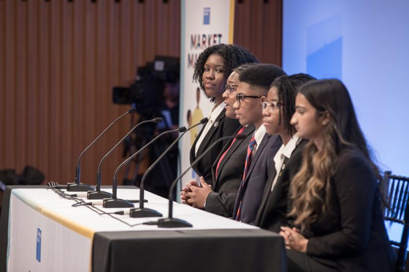 Two male and three female students sit at a white table with microphones in front of them