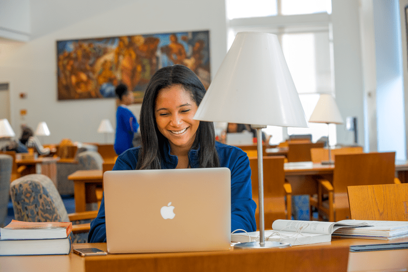 Black woman sits and smiles at her laptop while sitting in a library