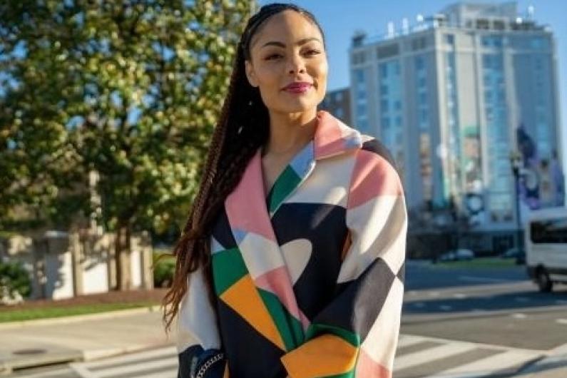 Woman with a colorful jacket on standing outside. In the background you can see trees and a building, as well as the street