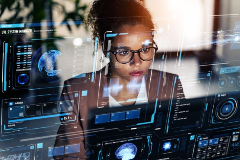 A woman with glasses sitting behind a computer with various kinds of digital text transparently around her face and the foreground, meant to represent aI