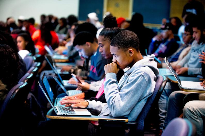 Students in a full university classroom with their laptops on the desks. The students are looking at their laptop screens intently. 