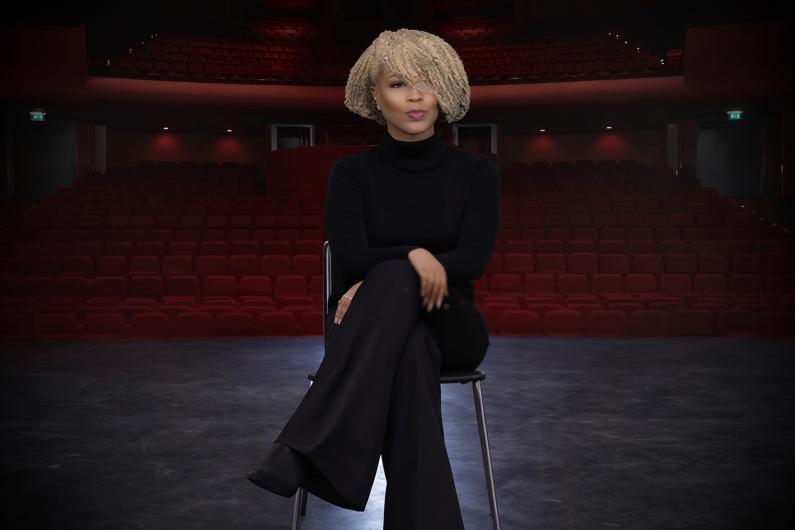 woman sitting in a chair while on a theatre stage. There are red theatre chairs behind her