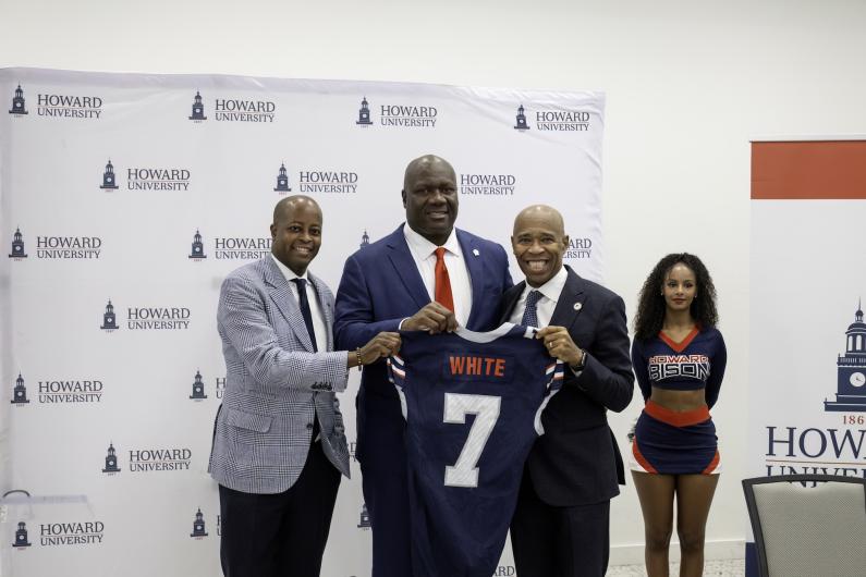 Howard University President Wayne A. I. Frederick, Coach Ted White and VP of Athletics Kery Davis pose holding Coach White's Howard football jersey. A Howard University cheerleader stands in the background.