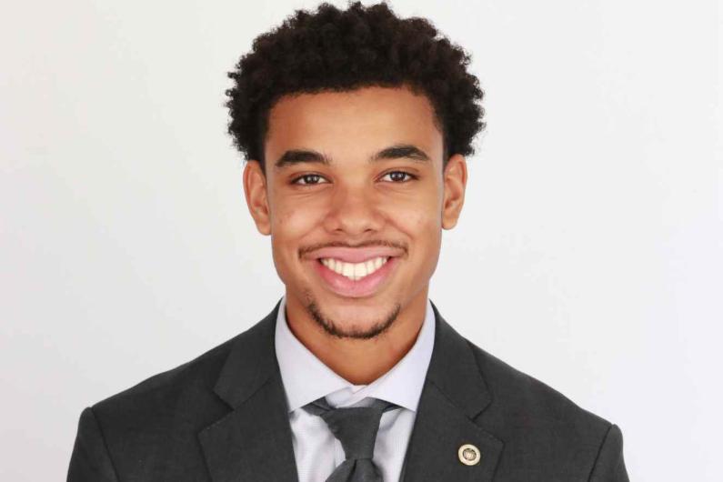 A headshot of a howard student, Stanley Scott, who is smiling at the camera. He is sitting in front of a white wall and has a dark suit on with a white shirt and a dark tie