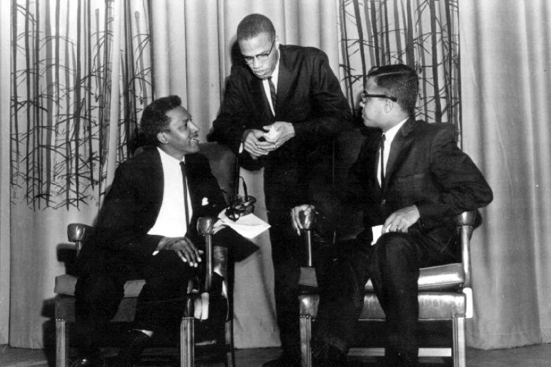 Malcolm X, Bayard Rustin and Michael Winston sit on stage at Howard University