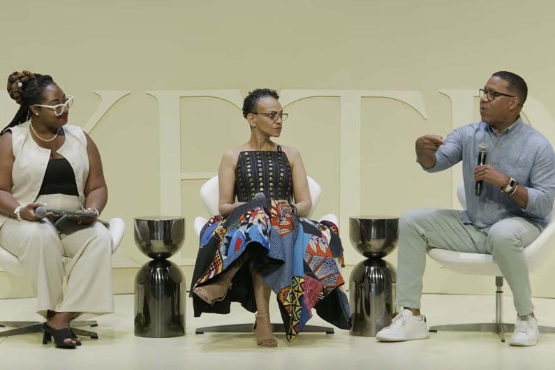 Three people sitting down on stage. Marcus johnson is on the far right, holding a mic and gesturing while talking