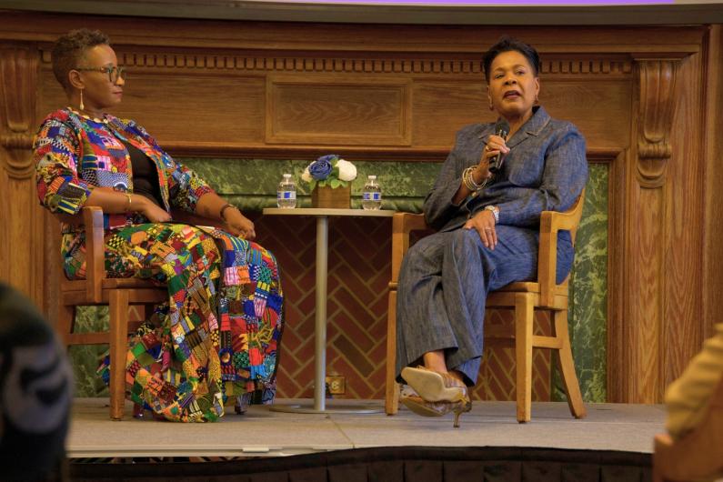 Two women sitting on a stage with microphones in their hands. There is a small table in between them with two small water bottles on top
