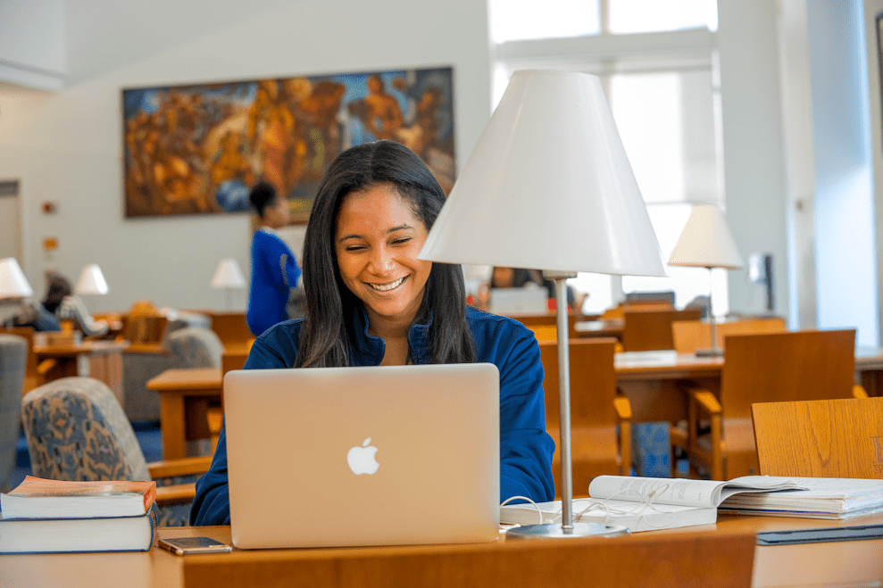 Black woman sits and smiles at her laptop while sitting in a library