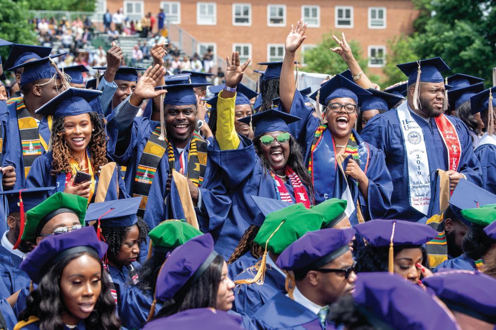 A group of Black students dressed in graduation regalia cheer on the Howard University Yard during a commencement ceremony