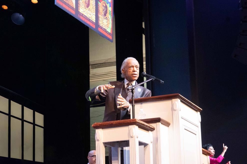 Man standing behind a pulpit-like podium, gesturing with his hands as he talks. There is a woman on the far right doing ASL alongside him