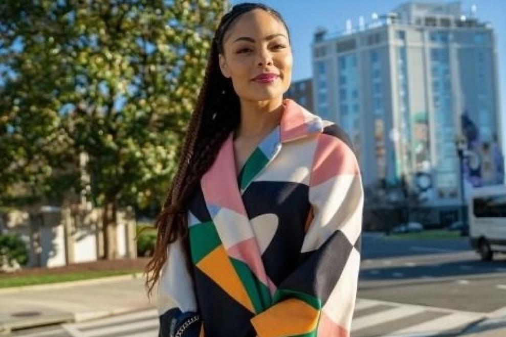 Woman with a colorful jacket on standing outside. In the background you can see trees and a building, as well as the street