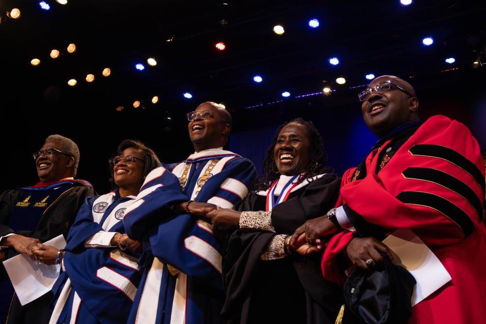Five Howard University leadership standing side by side, locking hands while looking out into a crowd for the university's convocation. They are all wearing university regalia robes of different colors such as red, black, and blue
