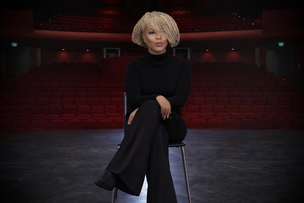 woman sitting in a chair while on a theatre stage. There are red theatre chairs behind her