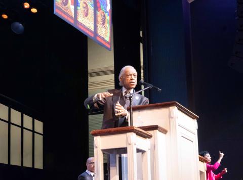 Man standing behind a pulpit-like podium, gesturing with his hands as he talks. There is a woman on the far right doing ASL alongside him