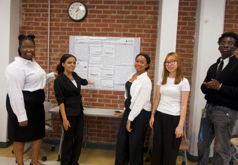 Five students smiling at the camera while standing in a classroom. Two students on the left are pointing to a poster board that displays research findings