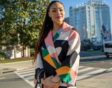 Woman with a colorful jacket on standing outside. In the background you can see trees and a building, as well as the street