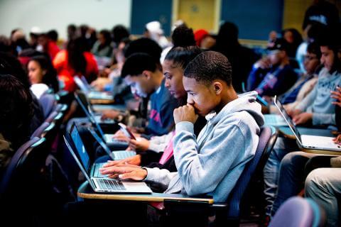 Students in a full university classroom with their laptops on the desks. The students are looking at their laptop screens intently. 