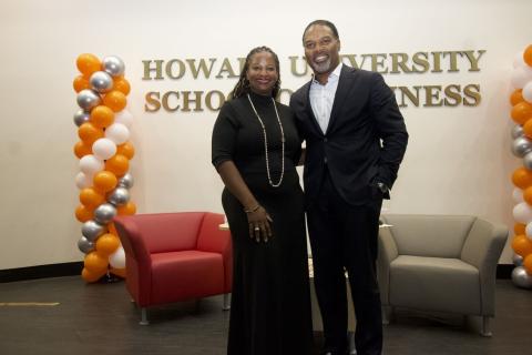 Two people standing side by side in front of a sign that says Howard University School of Business. There is a multi ballon structure in different colors on the right and left side of the image