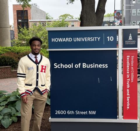 Graduating senior Joshua Taylor standS beside the Howard University School of Business sign wearing a cream colored Howard University cardigan