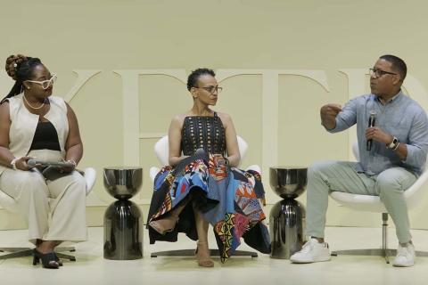 Three people sitting down on stage. Marcus johnson is on the far right, holding a mic and gesturing while talking