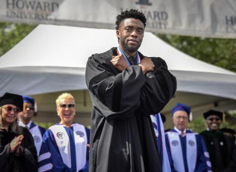 Chadwick Boseman doing the Wakanda Salute pose on stage. He has his arms crossed on his chest. There are people standing behind him