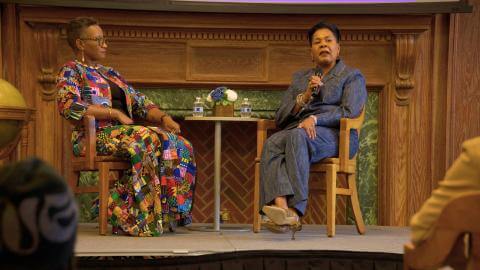 Two women sitting on a stage with microphones in their hands. There is a small table in between them with two small water bottles on top