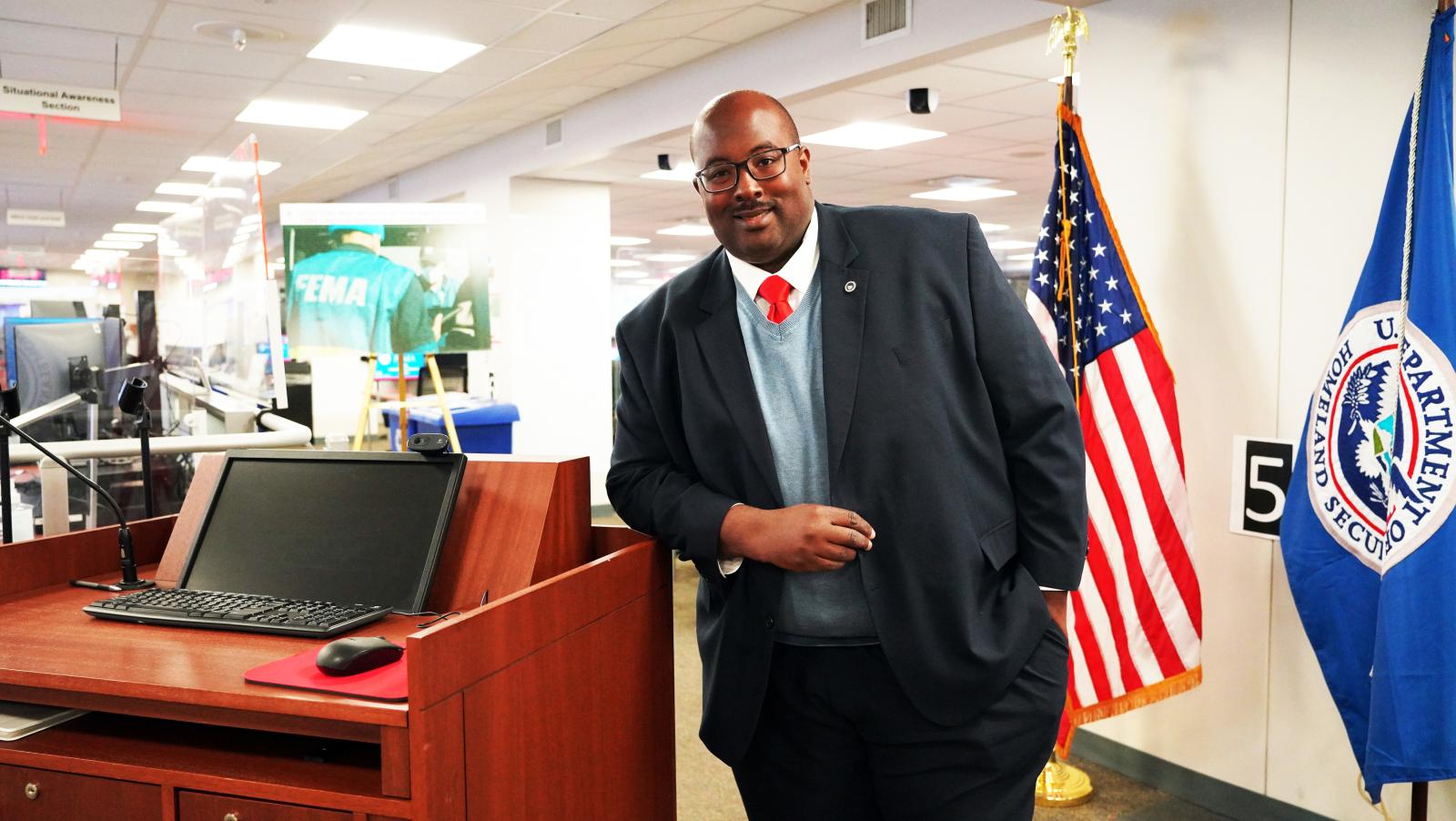 Marcus Coleman standing in suit inside FEMA offices with American flag
